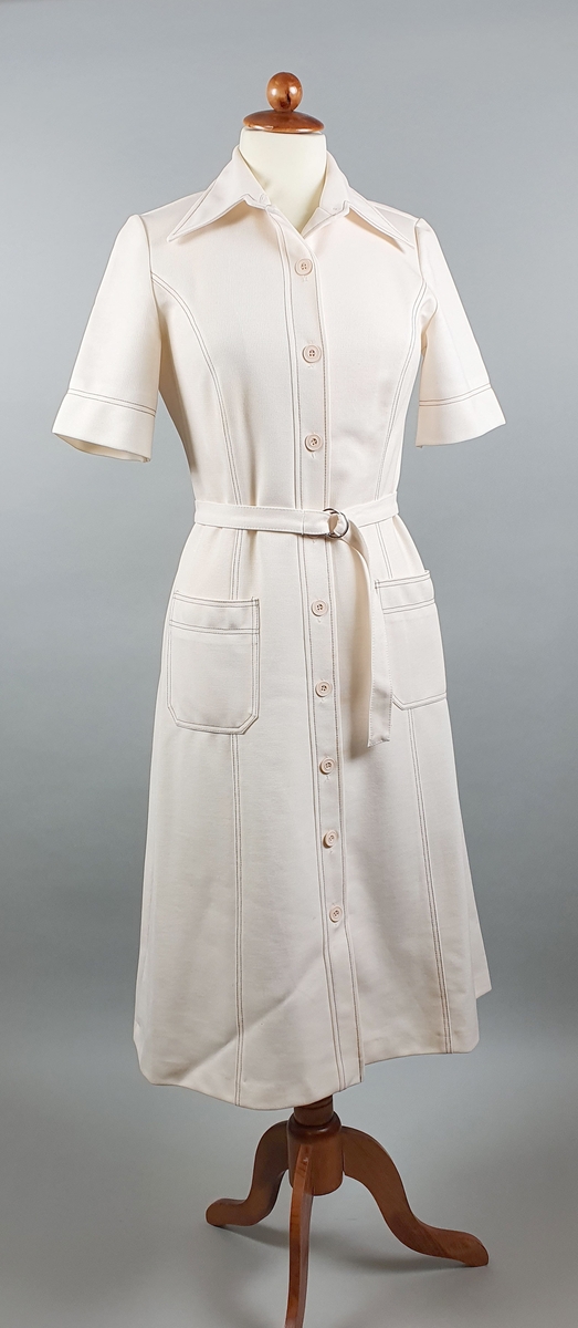 Hvit kjole med knapper foran, skjortekrage, påsydde lommer og belte. Brune dekorsømmer.