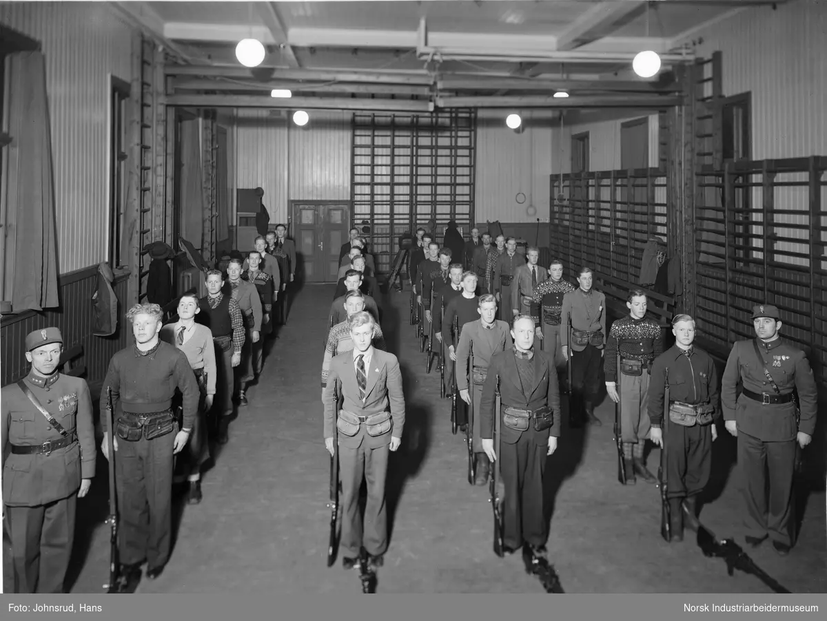 Frivillig militæropplæring på Notodden. Menn stående med våpen i gymsal. To menn i uniform stående på begge sider.