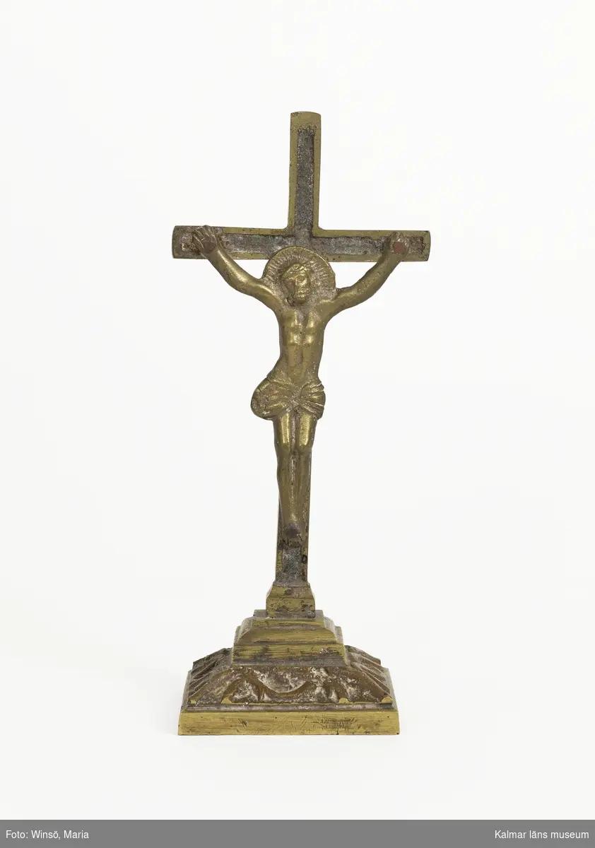 KLM 10337. Krucifix. Av brons, gjuten. På fot. Jesusfiguren är fastnitad i korset.