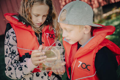 Foto av to barn med redningsvest på som kikker i en lupeboks etter vann insekter
