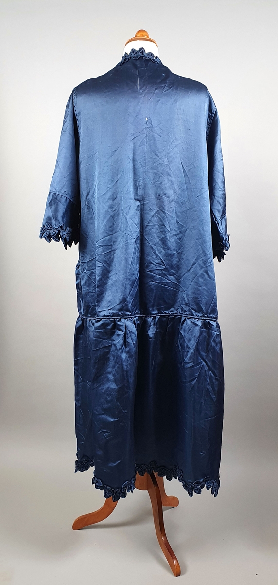 Mørkeblå kjole (morgenkåpe) av silke med rysjekanter nederst, langs åpningen foran og ytterst på ermene. Påsydde lommer med rysjekant og blomsterdekor.