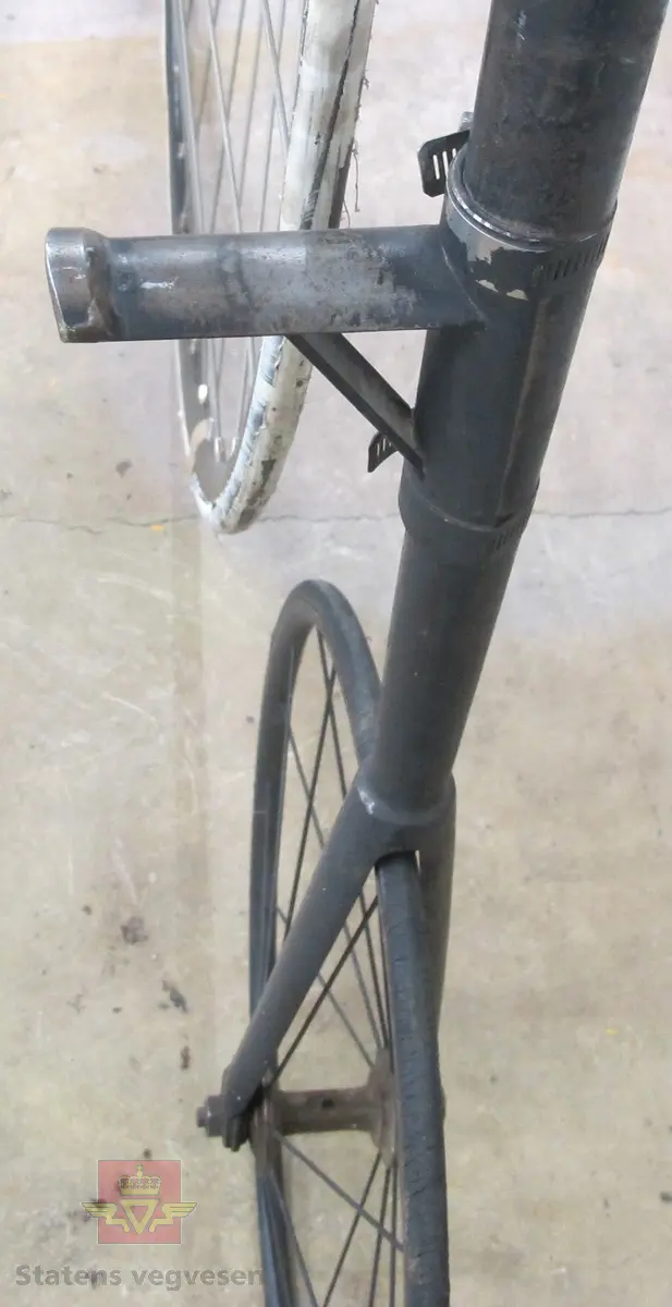 Sortlakkert velosiped med stort forhjul og lite bakhjul. Pedalene er montert direkte på fornavet og setet er plassert rett over. Hele sykkelen er av metall untatt håndtakene og dekk. Håndtakene er av tre og dekken av gummi.