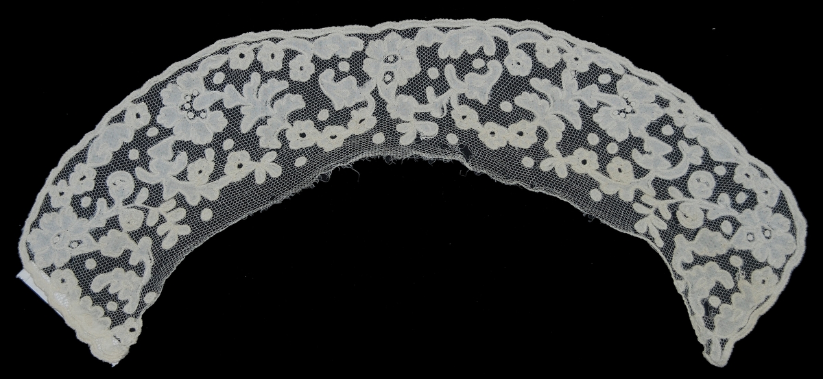 Krage bestående av rundad tyllremsa prydd med applikationsbroderi av tunn vit linnelärft fastsydd med vitt lingarn med kedjestygn. Mönstret bildar blom- och bladmotiv.