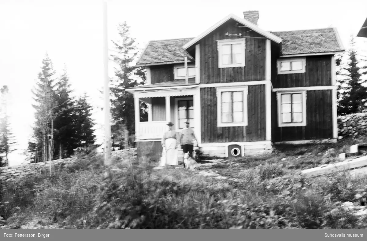 En kvinna, en man och en hund framför ett bostadshus vid Bergavägen 29, Njurunda. I det huset bosätter sig senare fotografen Birger Pettersson.