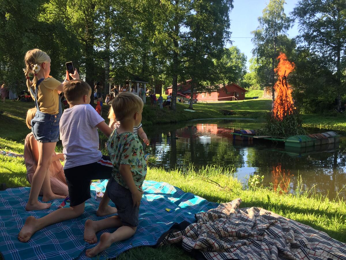 Tre barn i forgrunnen med ryggen til, ser på et stort, fint og flammende sankthansbål ute på et tjern på Eiktunet Friluftsmuseum, Gjøvik. Sol, grønne plener og store trær rundt vannet.