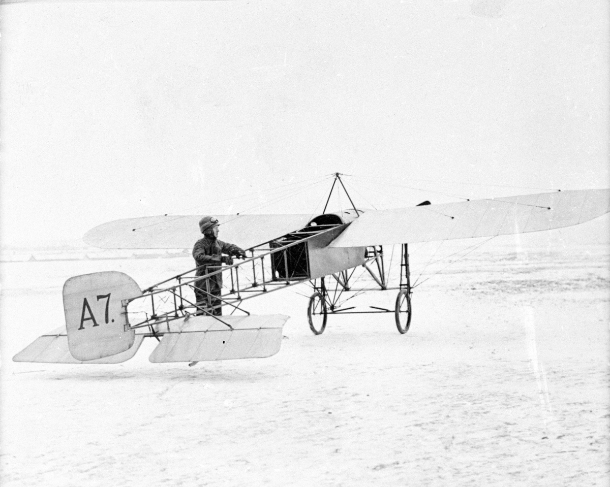 Flygplan typ Thulin A, märkt nummer A 7, står på ett flygfält vid Thulins flygskola, vintertid omkring 1917. Vid flygplanet står flyglärare Axel Lindh.
