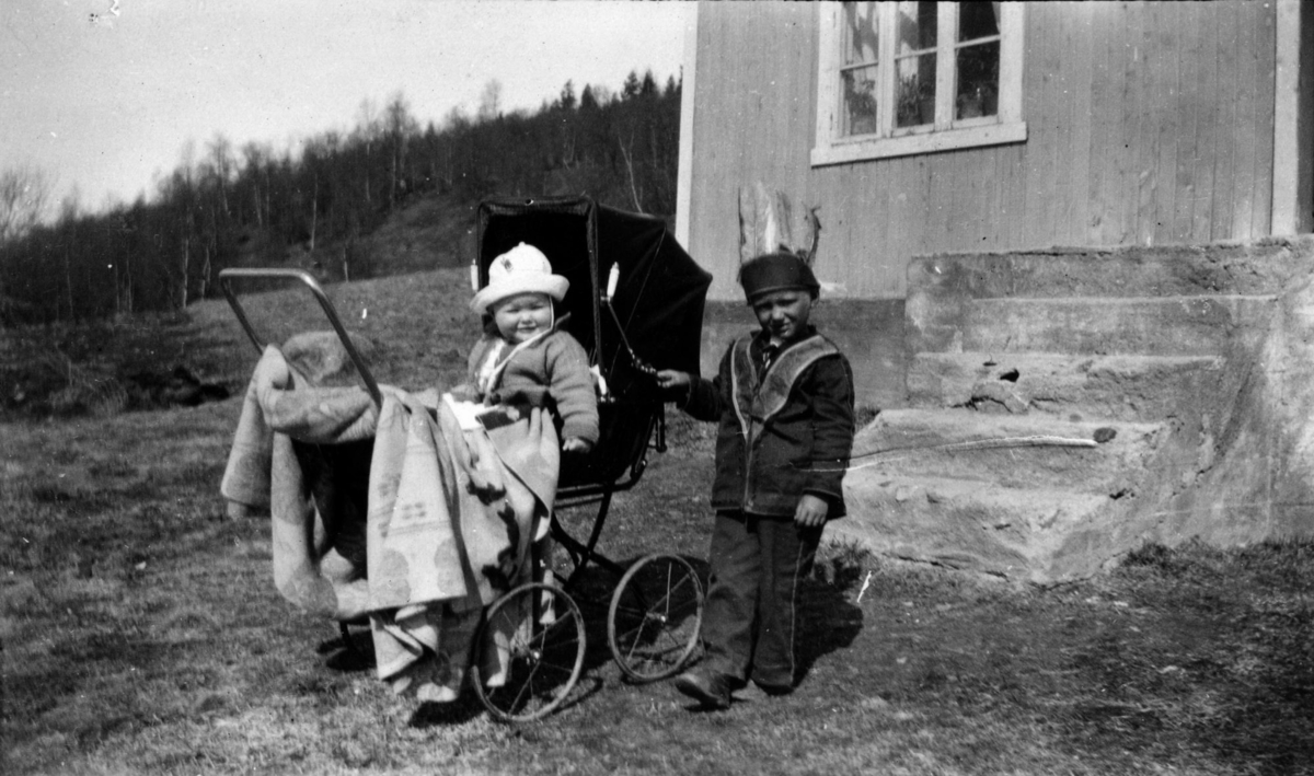 To små barn foran hovedstua på gården Helstad i Målselv i 1930. Svanhild Myhre sitter i barnevogn, men Hans Wiesner står ved siden av. Hans har kledd seg ut med fjærbekledning på hodet, leker han indianer?