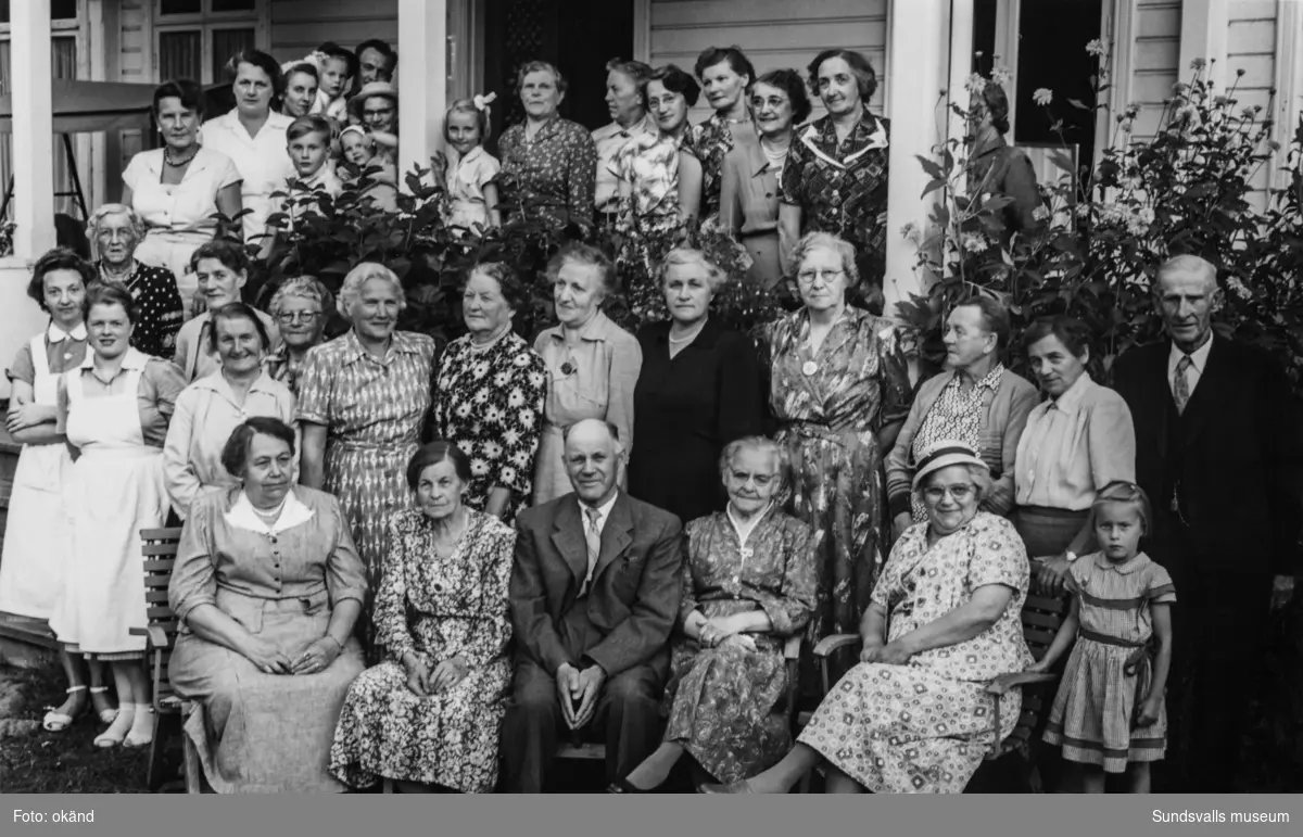 Gruppbild med 36 personer, övervägande kvinnor samt några män och barn, som står och sitter framför verandan till ett hus. Ur en samling från Svartvik.