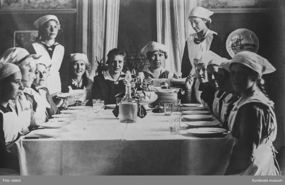 15-talet flickor med vita förkläden och hucklen samt en kvinna (lärarinnan?) runt ett dukat matbord. På bildens baksida står: Skolkök i Skönvik. Ur en samling som tillhört Ada Olsson, Målås, Skön.