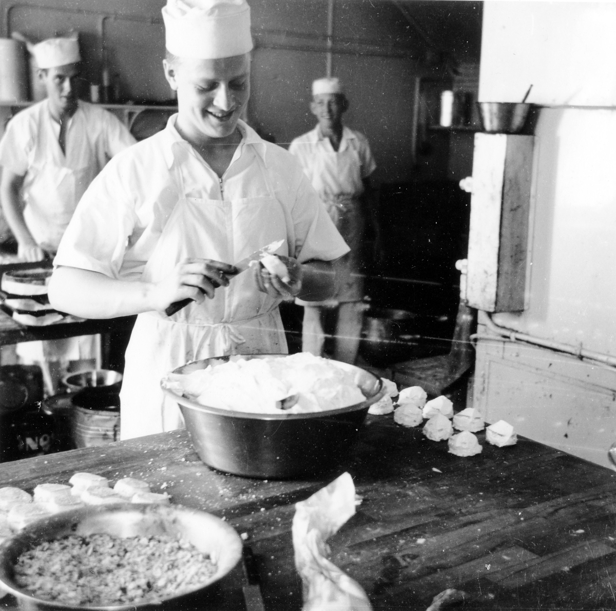 3 män i vita arbetskläder, förkläden och mössor arbetar i köket på Gustavs konditori, kv Gustaf vid Stora torget. 
Innehavaren av konditoriet hette Gustav Johansson enl adresskalendern 1950.