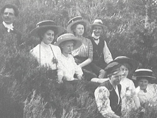 Utflykt. Ett sällskap män och hattbeklädda kvinnor sitter i en skogsbacke.