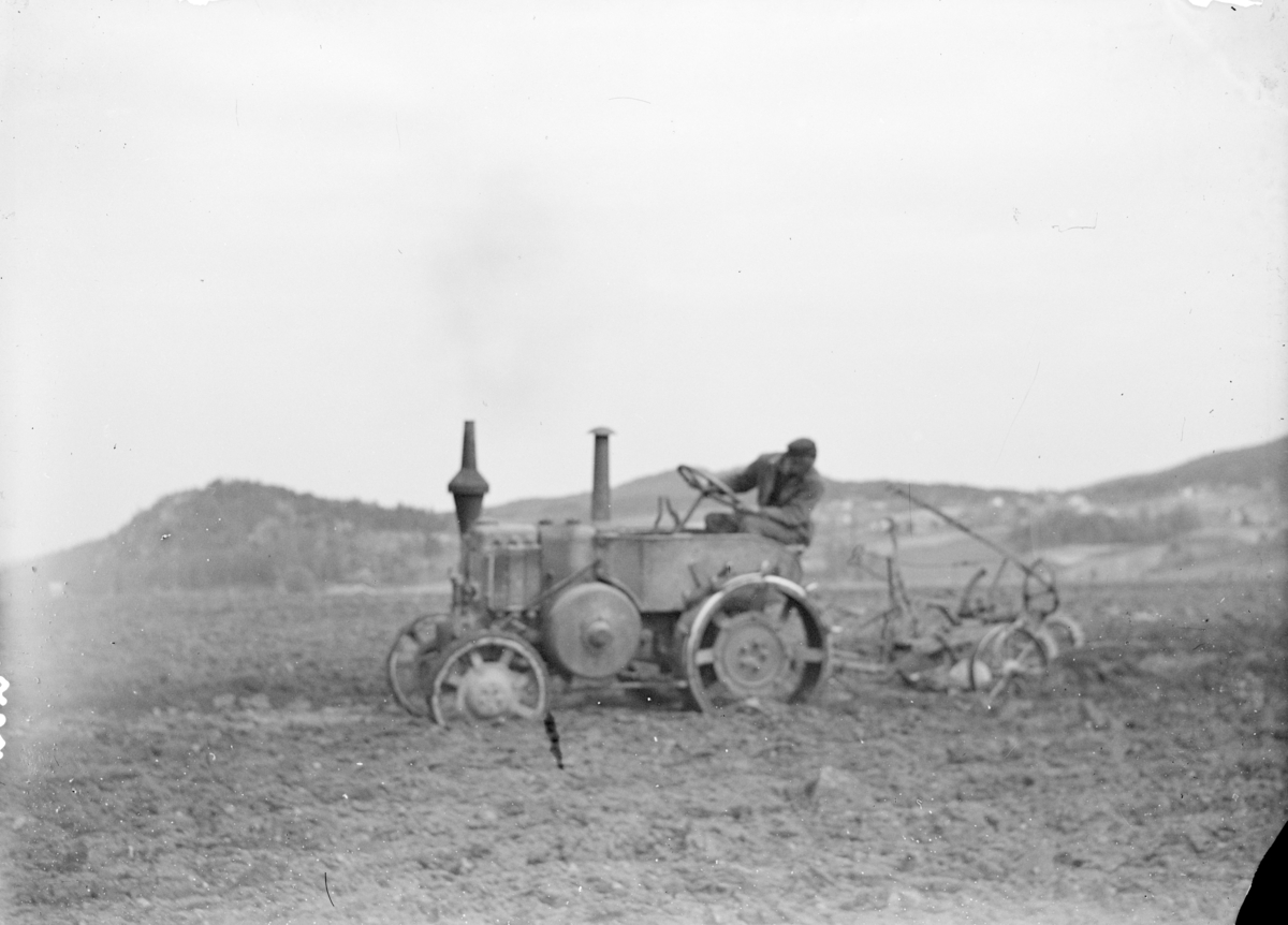 Fotografi av en traktor som trekker en plog på et jorde. Tekst fra Teigens katalog: "Svelviks Historie, Svelviks museum etc. se også: Drammens Tidende, se også: Hurum Bygdebok 4938"