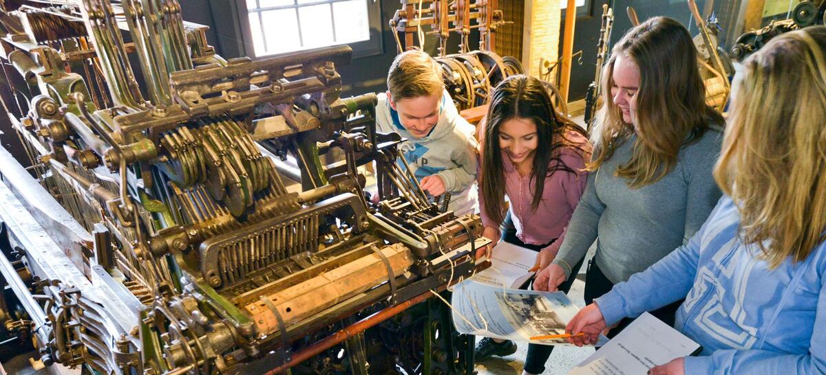 Bildet viser fire unge jenter som holder i penn og papir  ved siden av en maskin.