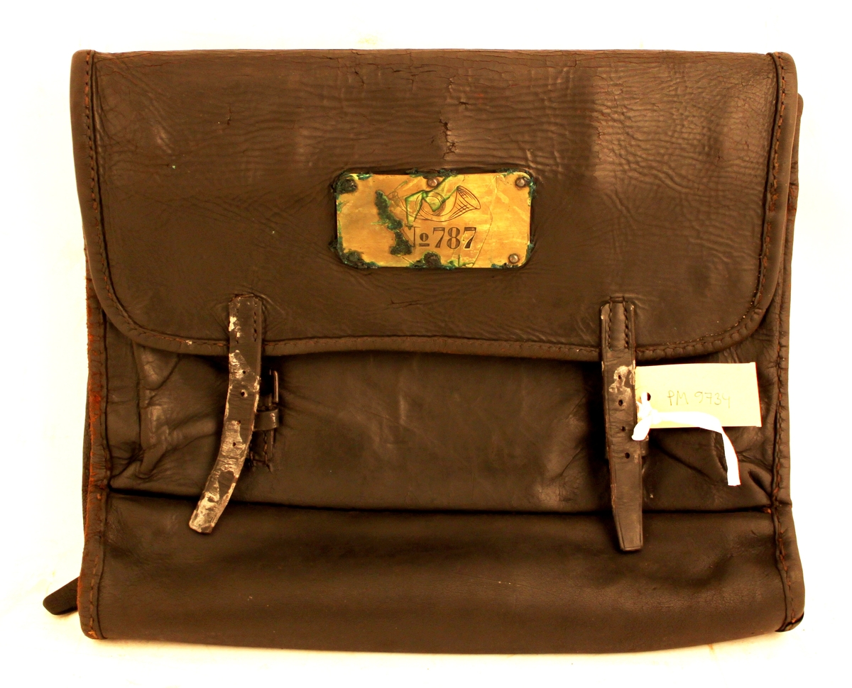 Väska av svärtat läder med klaff och två remmar och hålspännen. Låses med järnten (saknas). På klaffen en mässingsbricka märkt med "No 787".