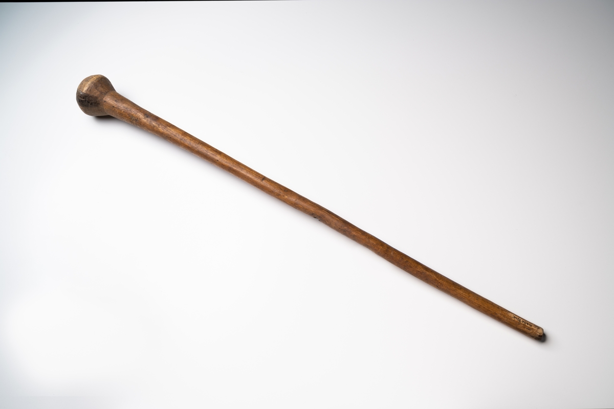 Mortelstöt av trä i form av en lång käpp med en klumpformad avslutning i ena änden.

Se vidare Historik.