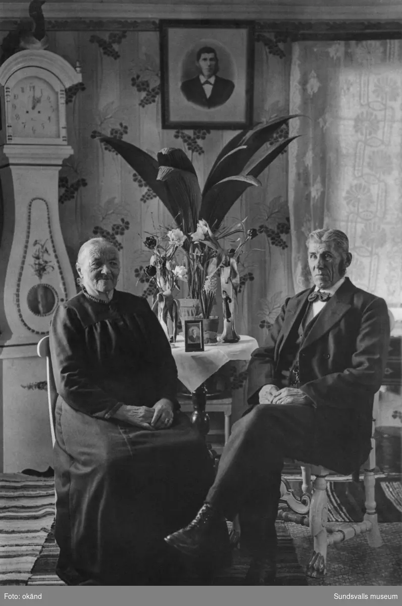 Porträttbild av ett äldre par i hemmiljö. Blommor på bordet, golvur och porträttbilder på bord och vägg. Ur en samling som tillhört Viklander i Backås, Hässjö.