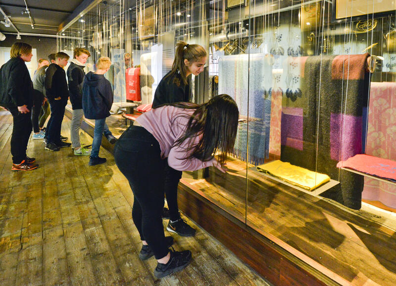 Bildet viser ungdommer som ser på en utstilling av tepper som henger i et avlangt stort glassmonter