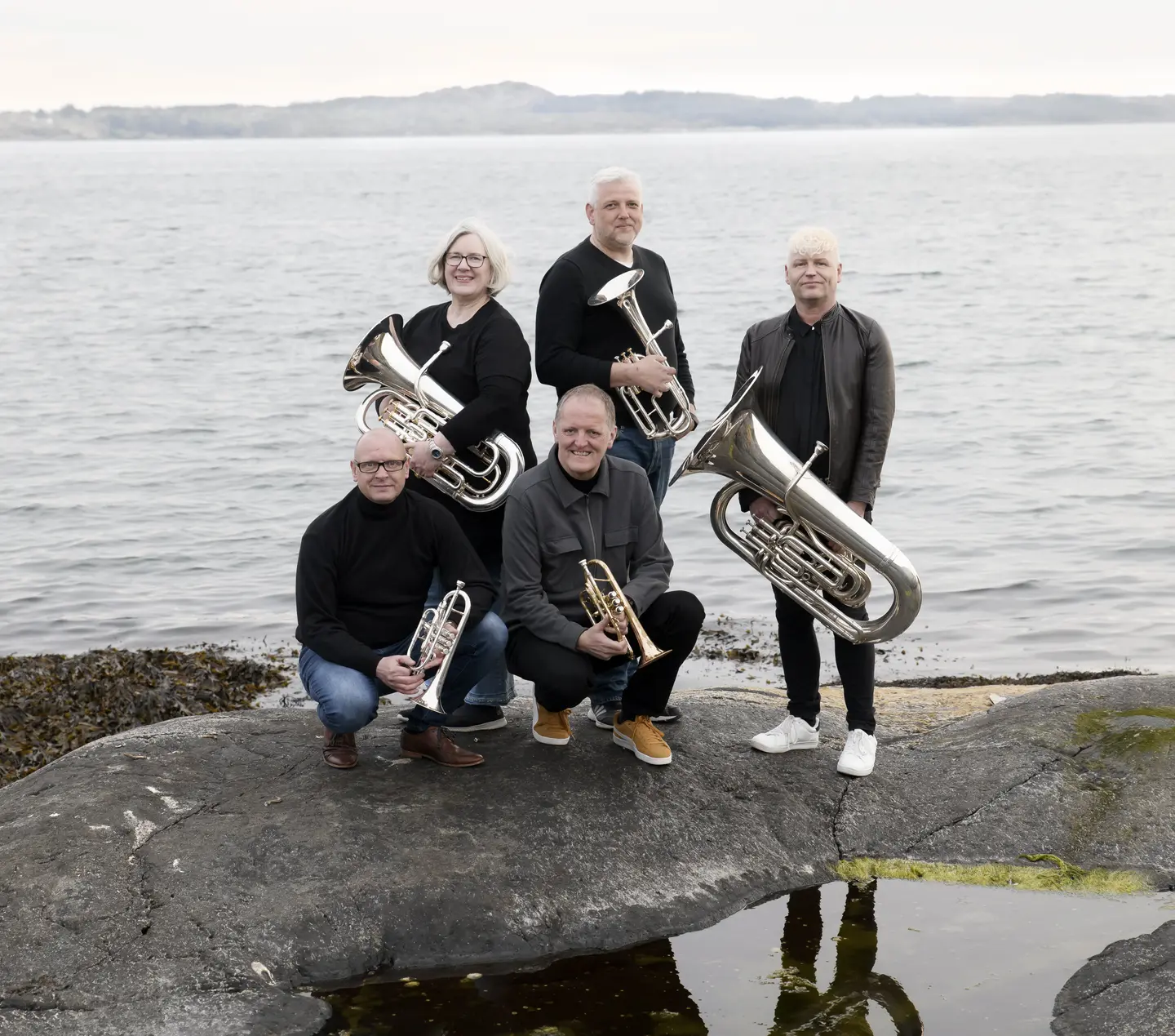 Fem brassmusikanter med instrumenter på svaberg i sjøkanten