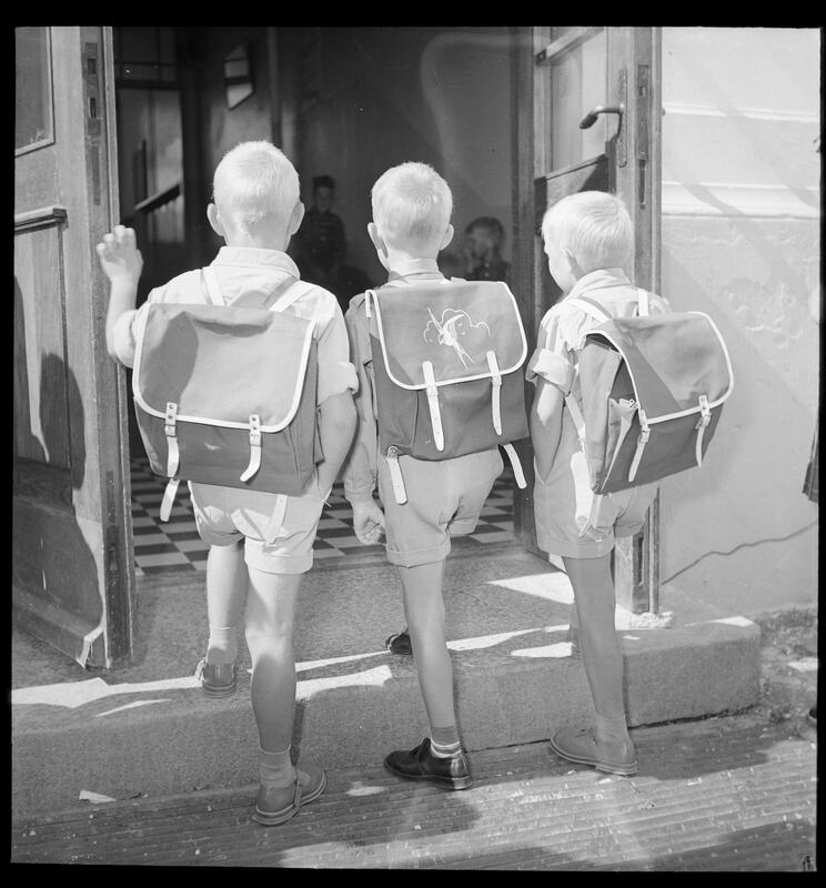 Sort-hvit-bildet viser ryggene til tre små gutter i shorts og kortermet genser og helt like skolesekker. De går opp et trinn og inn en dør.