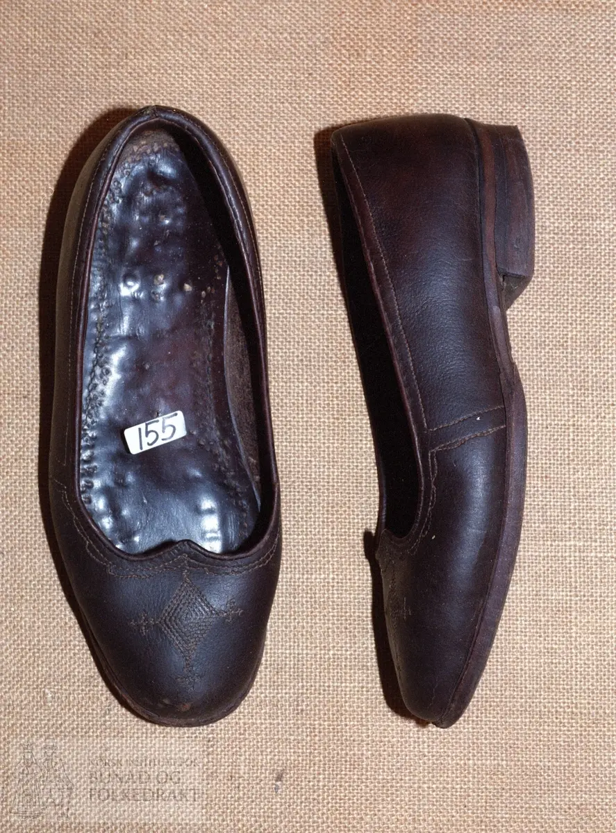 Laga eigar av skomaker Solum frå Lyngdal i 1949.  -  Pumpsforma sko laga av brunsvart lær. Sydd dekor framme på skotuppen med skråstilt rute med krossar i 3 hjørne. Stikningar etter kantane. Lærsolar under.