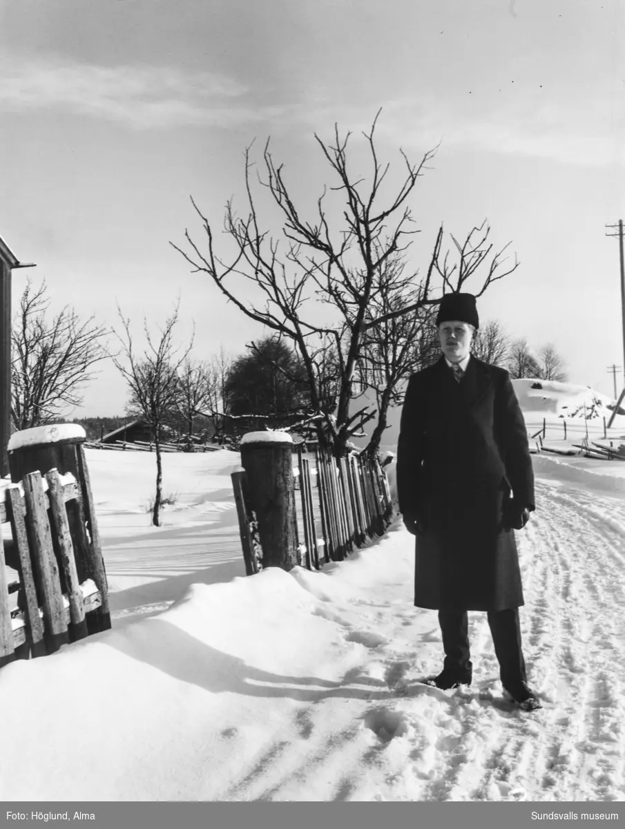 En man med rock och vintermössa står på vägen intill ett staket i ett vintrigt landskap.