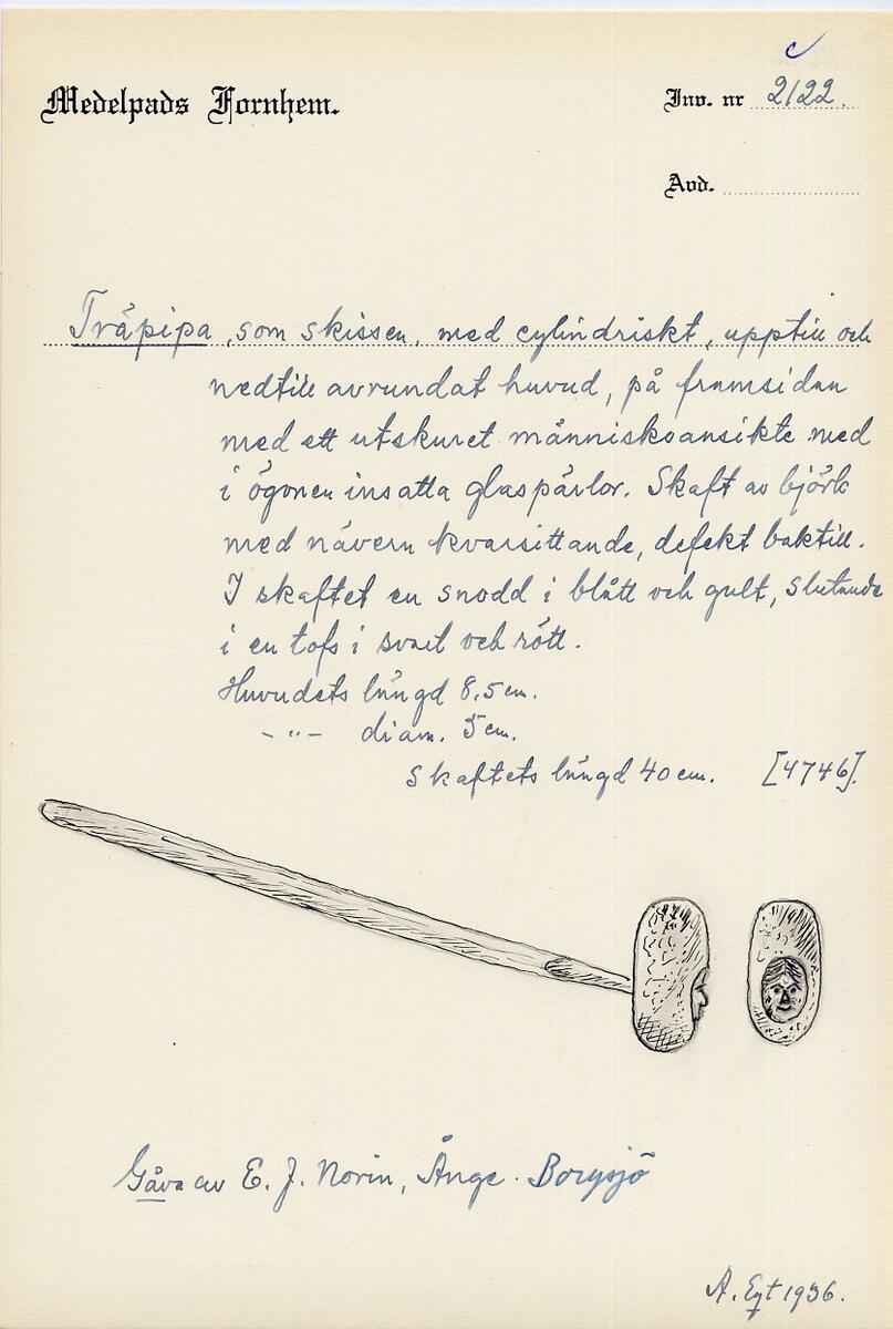Träpipa, med cylindriskt huvud, avrundat upptill och nedtill, på framsidan med ett utskuret människoansikte med ögon av glaspärlor. Rakt skaft av björk med nävern kvarsittande, mångfärgad. Skaftet med snodd i blått och gult, slutar med  en tofs i svart och rött, skaft saknas. Tillverkad 1850. Gåva av E J Norin, Ånge. 
