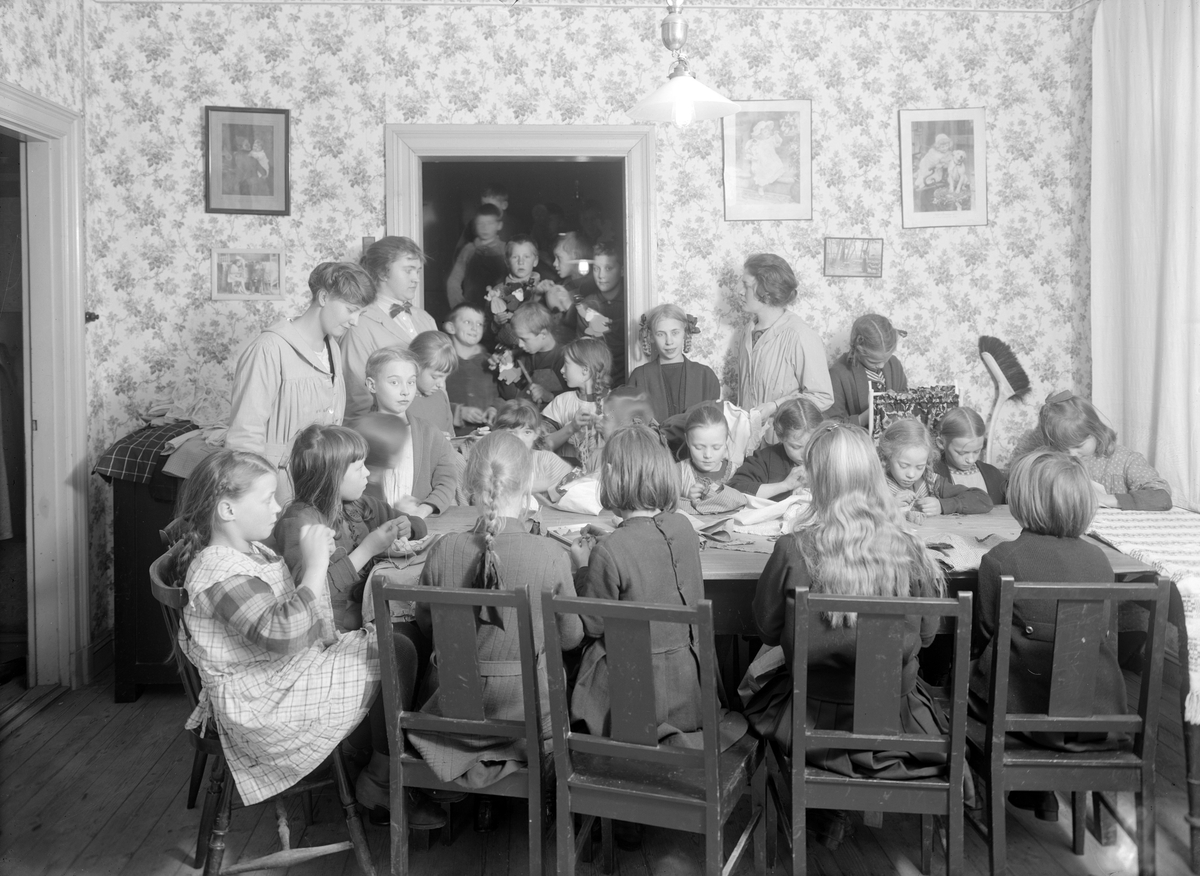 Interiör från Linköpings Arbetsstuga 1923. Bakom benämningen kunde varianter förekommma men i de flesta fall var det en praktisk skola, där fattiga barn undervisades i för deras färdighet lämpligt handarbete. Barnen erbjöds även åtminstone ett mål mat. Ändamålet var att motverka sysslolöshet och fattigdom. I Linköping låg vid tiden arbetsstugan på gården till Nygatan 43 och verksamheten förestods av fröken Annie Svensson. Möjligtvis är det henne vi ser invid dörröppningen. Bilden visar i övrigt flickornas avdelning som bokstavligen hölls skild från pojkarnas.