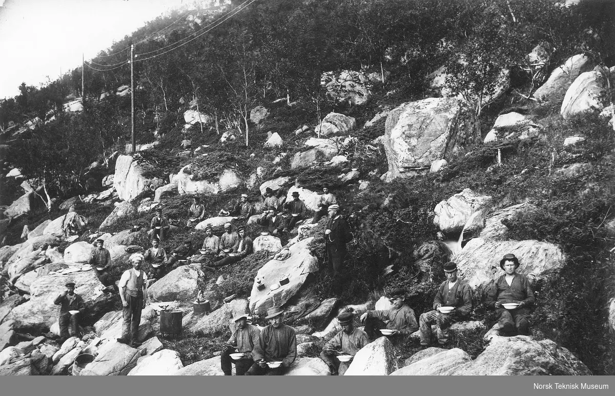 Kvinefossen : en flokk arbeidere har spisepause i fjellskråningen, kokken ved siden av en stor kjele