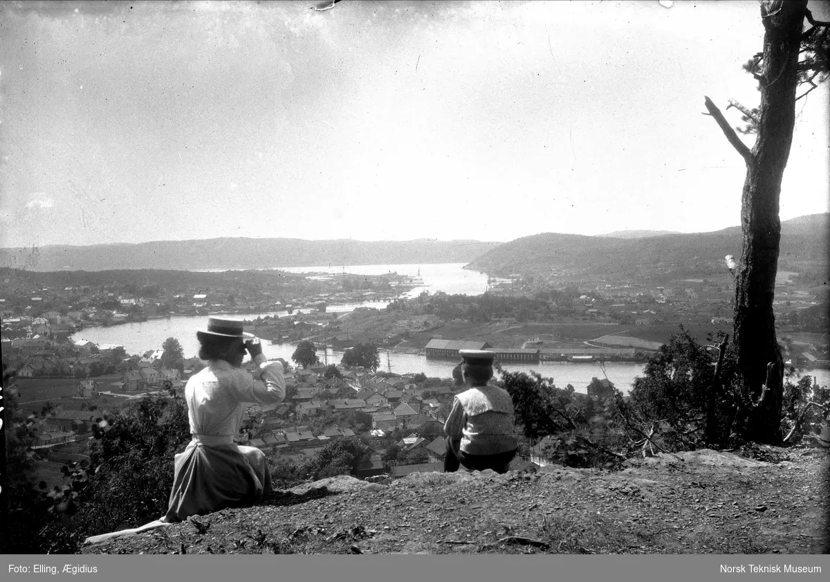Kvinne med hatt og gutt med matroslue skuer utover by ved elv