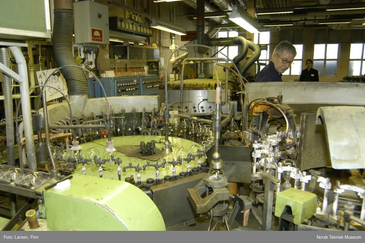 Osram-fabrikken i Drammen. Sammensmeltning av lampefot og kolbe overvåkes av mekaniker Lorang Axelson. I bakgrunnen Frode Weium