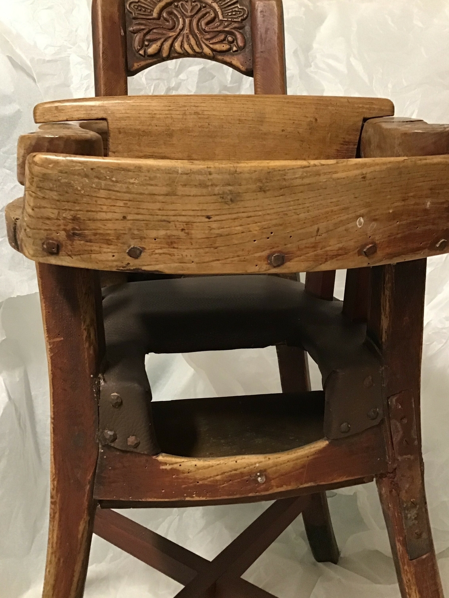 Barnestol  i tre med sete av skinn. Utovervendte stolføter med kryss. Svingbart bord i framkant av armlene. Utskore dekor i ryggparti.
