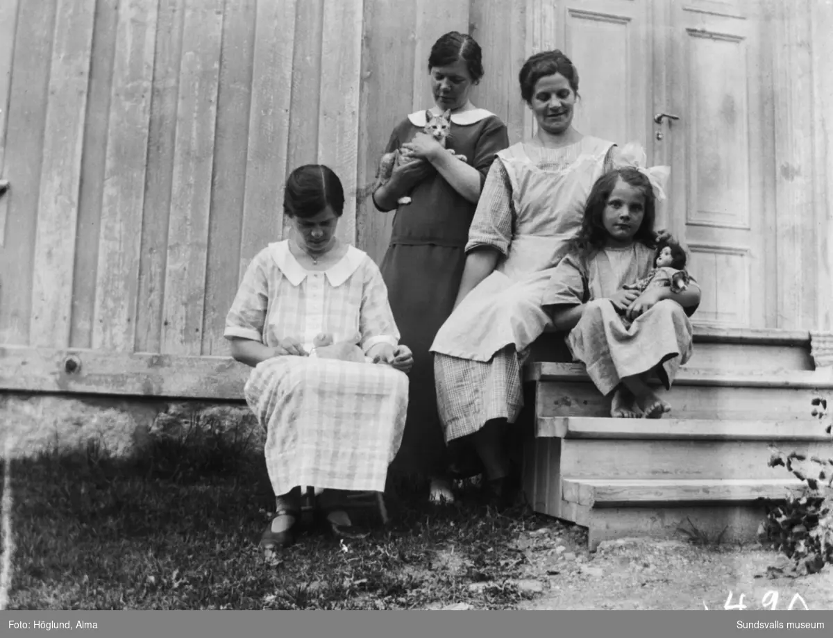 Tre kvinnor och en flicka på trappen till ett hus. Flickan har en docka i famnen och två av kvinnorna håller varsin katt.