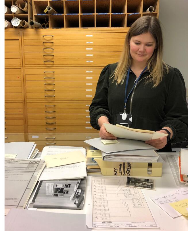 Bildet viser en ung kvinne som står ved et bord fylt av papirer. Hun ser ned på noe dokumenter hun holder i hendene. (Foto/Photo)