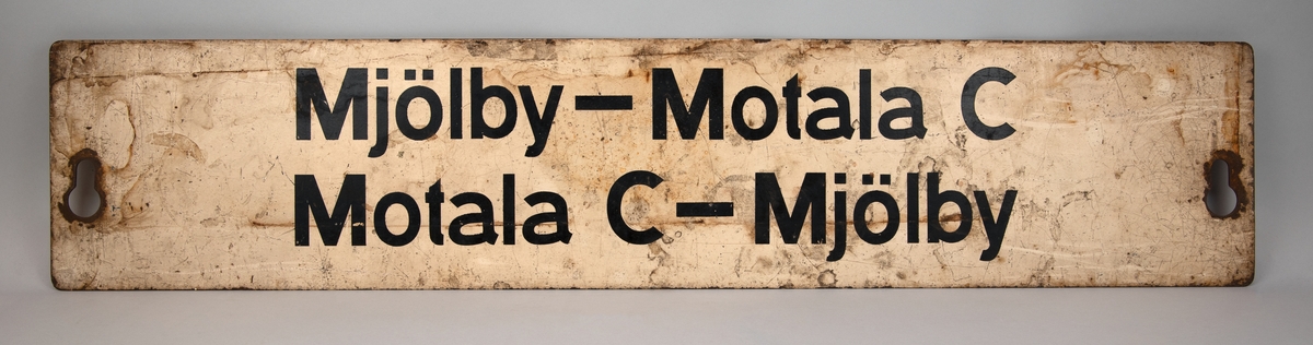 Rektangulär destinationsskylt av målad plåt. Vit botten med svart text, på ena sidan står det "Mjölby - Motala C", på andra sidan står det "Mjölby - Rönneshytta" och "My". Hål på kortsidorna för upphängning.