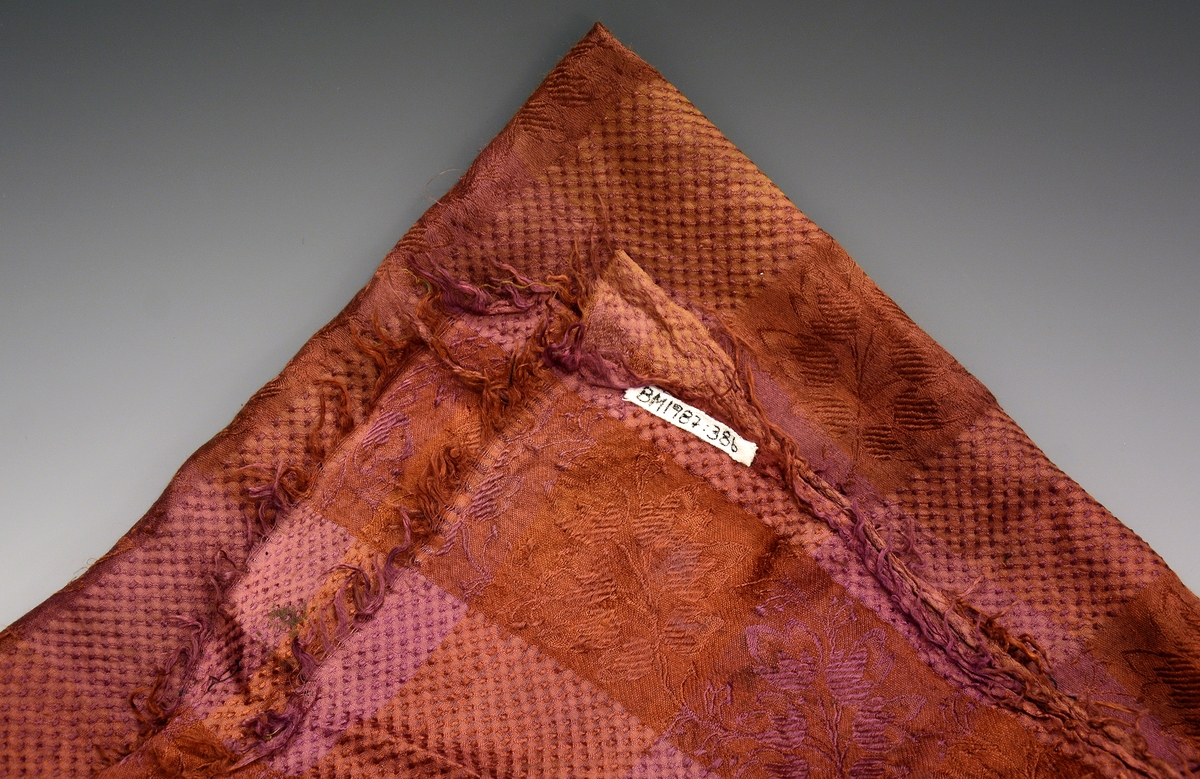 Tørkle, silke med innvevd mønster i fargene plommerødt og lys brunt. 7 cm. brede border, annenhver i blomsterranker og vaffelruter. Bordene danner kvadrater. Mål: 68 x 68 cm. Frynser rundt tørkleet.