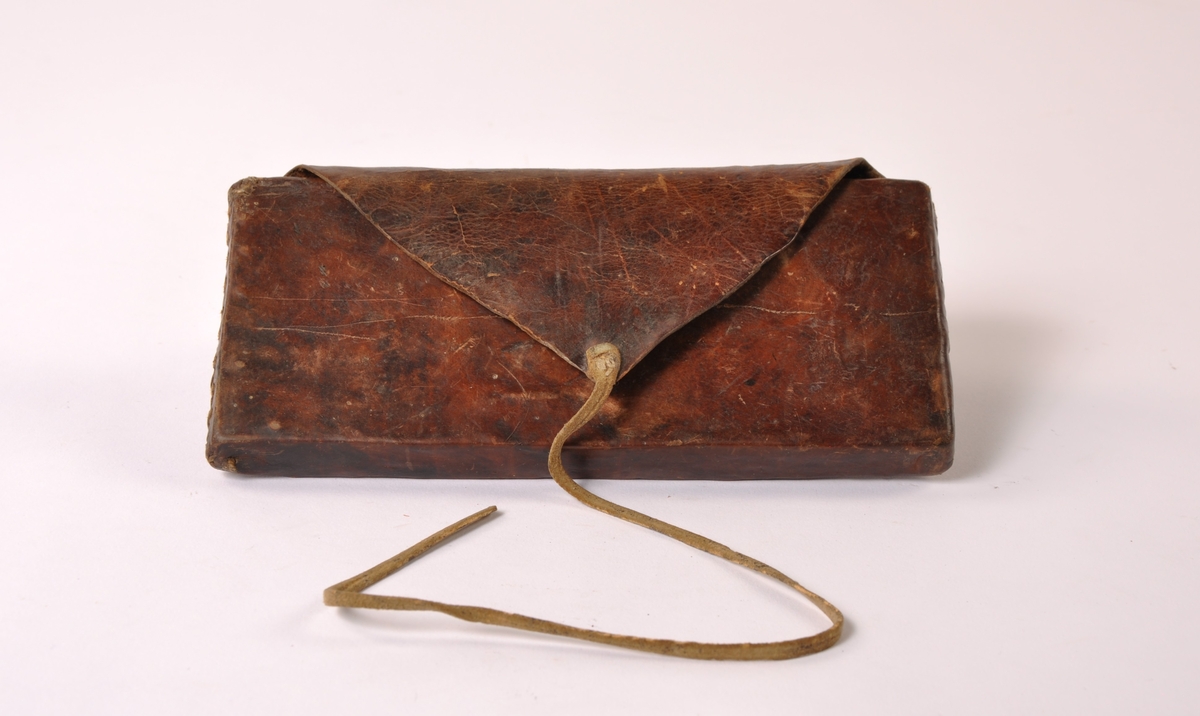 Lommebok i brunt lær, sydd sammen i sidene. Stivet med treverk innvendig og lærbånd som lås.
