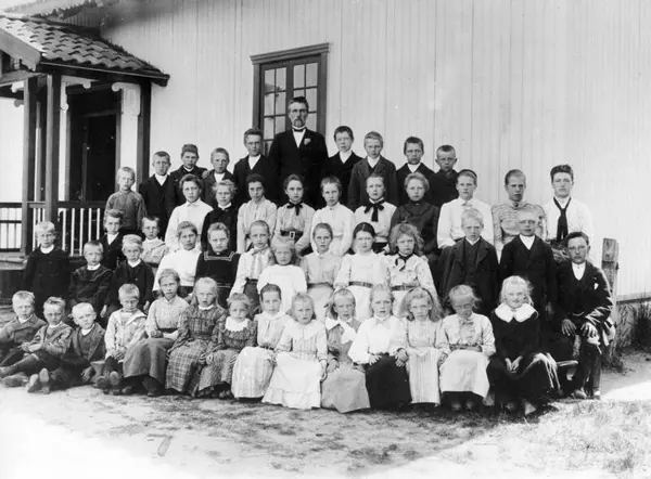 Fotografi i svart-hvitt av en skoleklasse og lærer Simensen. Elevene og læreren blir fotografert utenfor Skinnarbøl skole, som nå er skolemuseum.