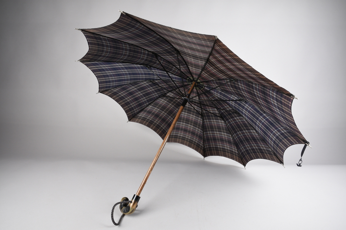 Paraply med 12 metallspilar. Stong i tre. Trekt med stoff i kunstfiber, rutemønster. Handtak i tre med hempe i tekstil til å ta inn på handa. Hempe for lukking av paraplyen.