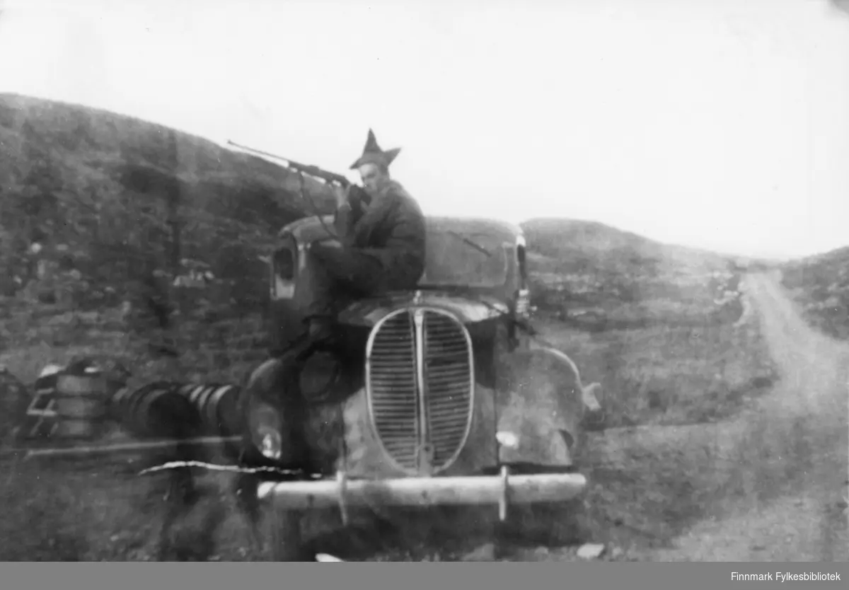 Sort hvitt fotografi av en mann med stjernelue, sittende på en Ford V8 1938 lastebil. Mannen holder et gevær i hendene. I bakgrunnen skimtes noen tønner. Bilen er parkert i veikanten ved en grusvei som forsvinner ut av bildet i høyre kant. Sted ukjent. Muligens Karasjok pga. stjernelua? 