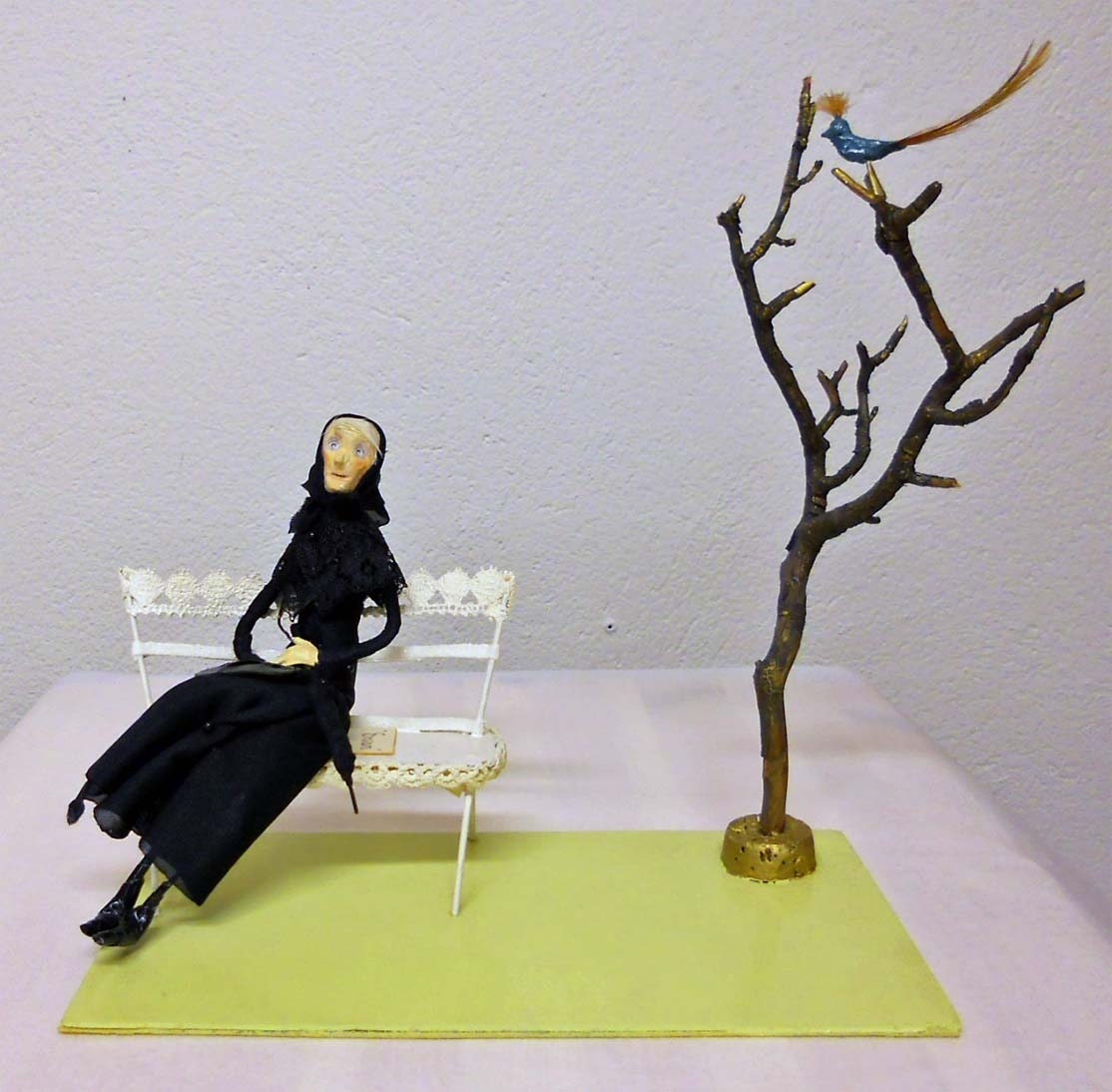 En gammal kvinna klädd i svart sitter på en vit bänk bredvid ett kalt träd. En blå fantasifågel sitter i trädet.