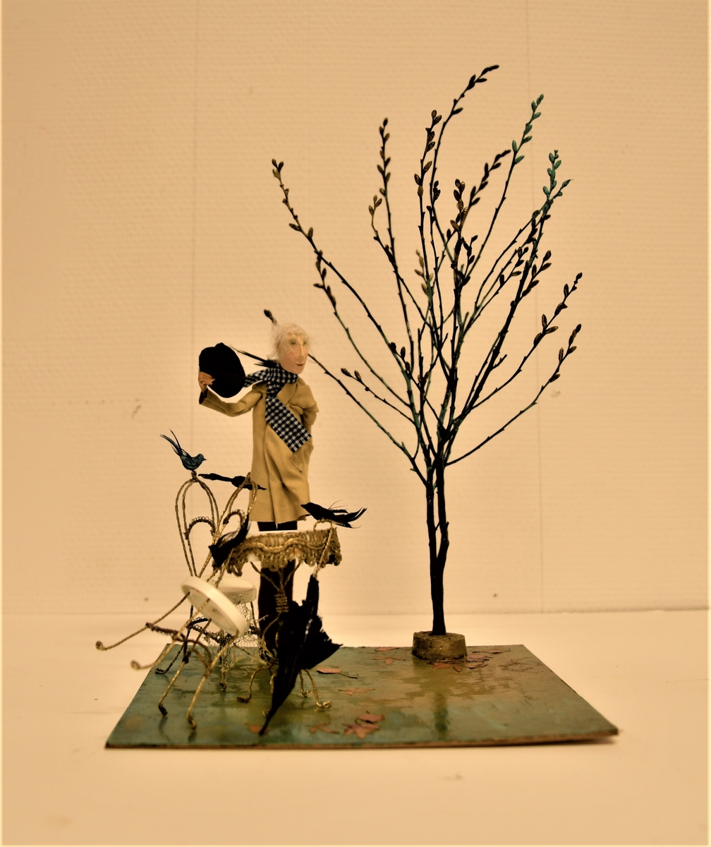 En vithårig herre i beige rock står vid ett kafébord med ett paraply och några fåglar. Han lyfter på hatten som till avsked. Ett kalt träd står bredvid.