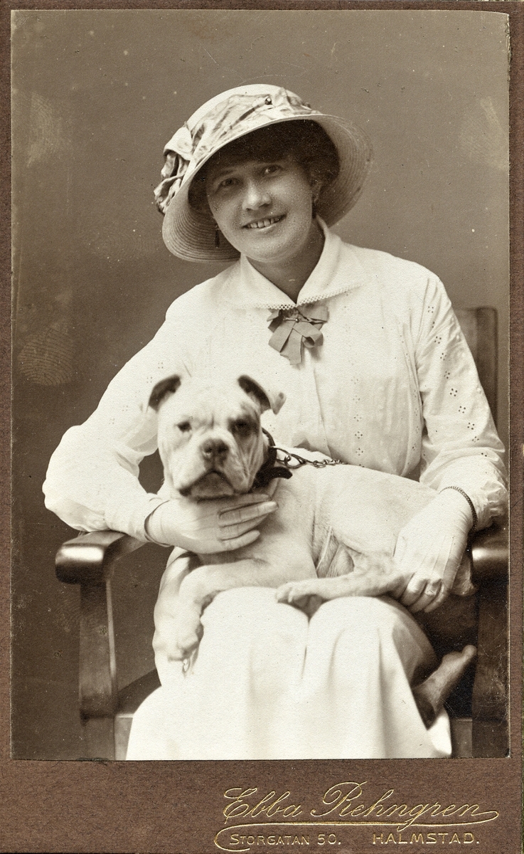 Ateljéporträtt av en okänd kvinna med hund i knät. Kvinnan bär hatt och handskar till dräkten, allt i ljus ton.