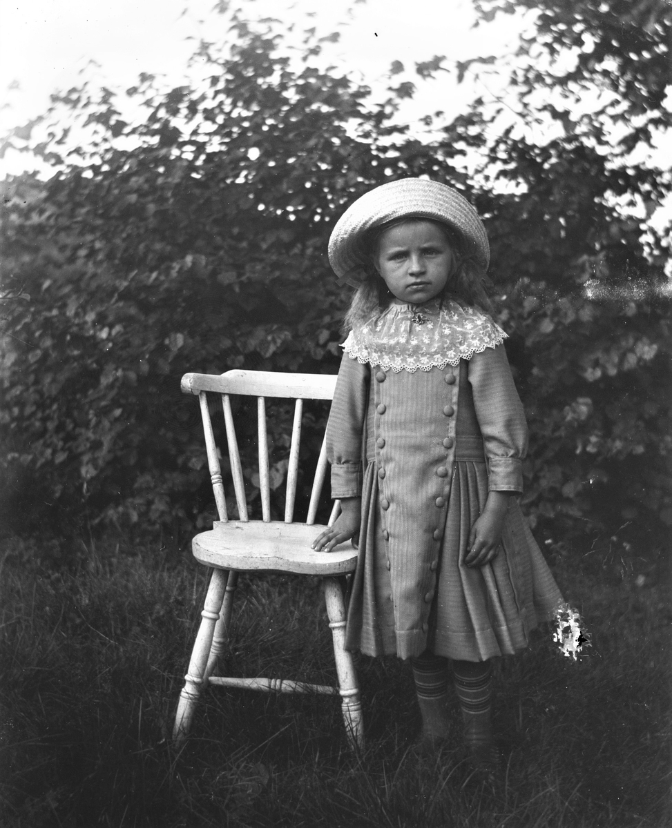 En liten flicka står utomhus vid en pinnstol. Hon bär stråhatt och en skir spetskrage på sin klänning med stråveck och klädda knappar. (Se hela familjen på bild nr GEB197)