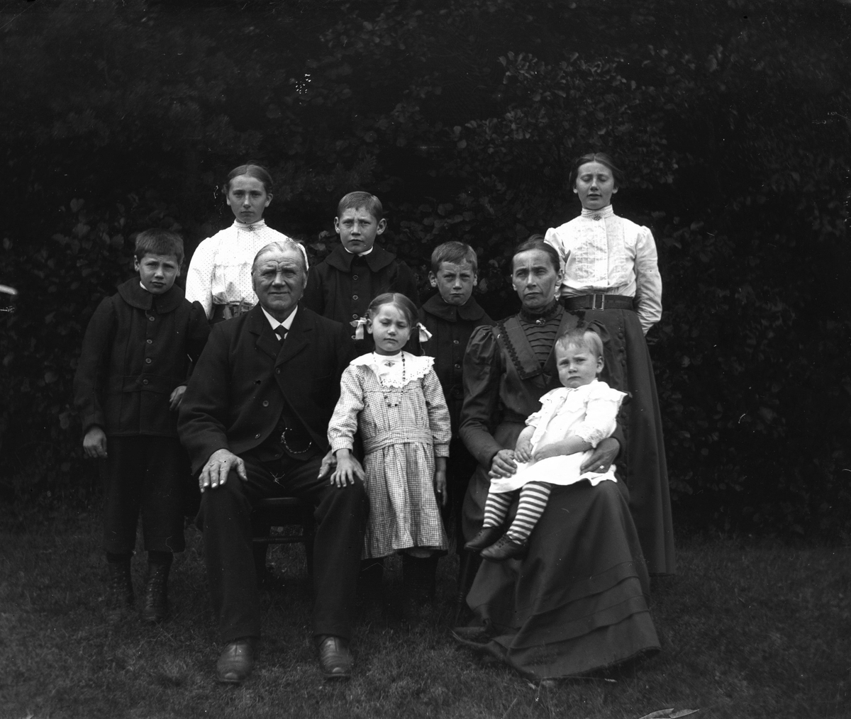 Familj med sju barn samlade utomhus för fotografering. Föräldrarna sitter och barnen står runtom. Tre pojkar har likadana jackor med rundade kragar. (Se även bildnr GEB184, GEB200)