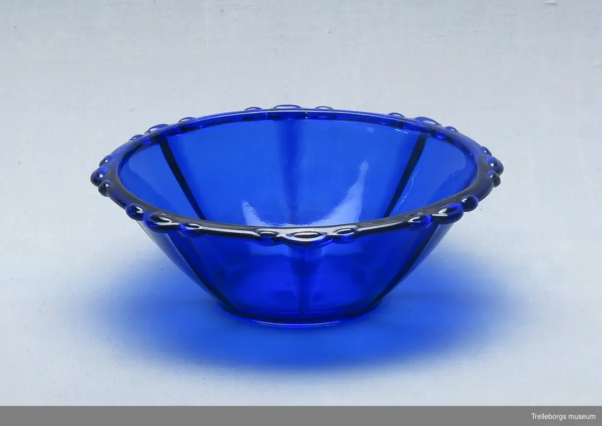 Skål i blått glas. Längs med kanten är ett utskjutande, runt mönster om 3 och 3.
