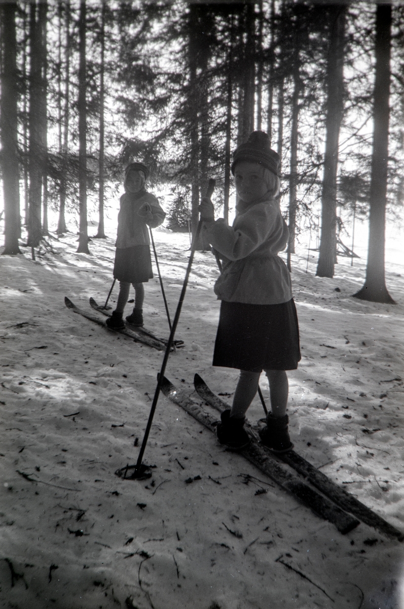 To jenter på ski fra Hol vestre i Vang til Nordre Arneberg i Romedal slutten av mars. Ikledd skjørt i anledning at de er på tur til en bursdag. Her på ski igjennom skogen. Jentene er Ragnhild Glærum (f: 1945) og Eli Vesaas (f: Glærum 1947). Fotograf Andrea Glærum (f: Gjestvang 1914)
