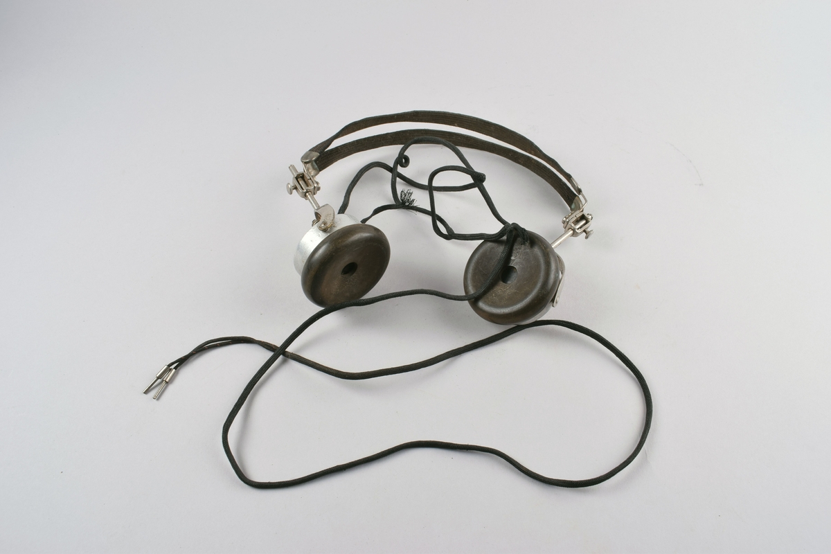 Høretelefoner forbundet med dobbel hodebøyle i metall trukket med tekstil. Ledning som splittes i to plugger nederst. Har vært brukt sammen med krystallapparat.