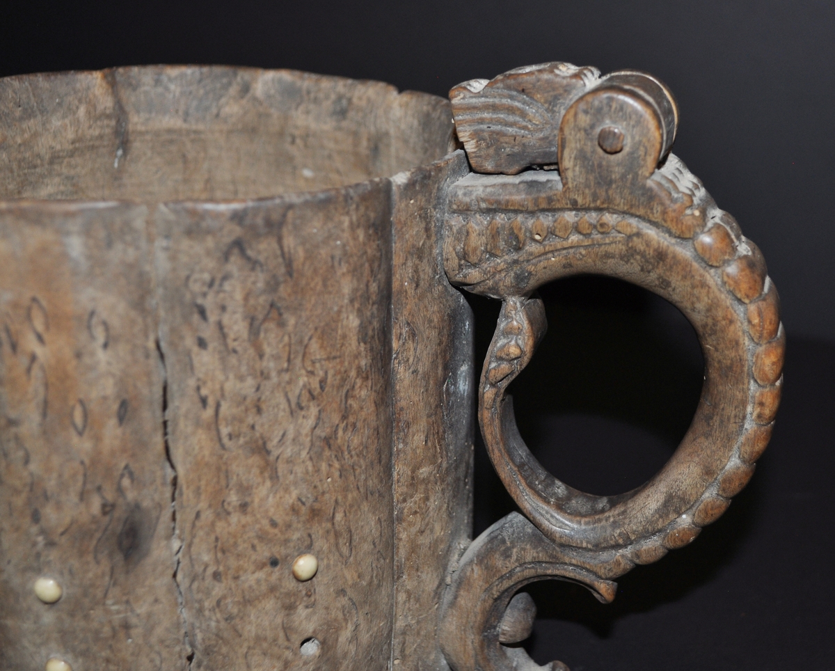 Dryckeskanna av masurbjörk med ett rikt utsirat handtag i form av en drake. Kannan är dekorerad med små runda benbitar. Barockform med välvda former utvändigt.
