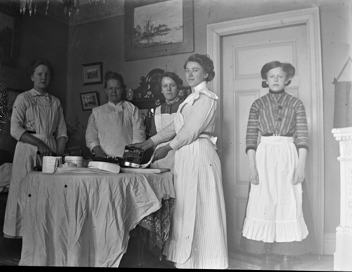 Fem kvinnor står vid ett runt bord i en salong och stryker löskragar. De bär förkläden så troligen är de tjänstefolk. På bordets tjocka duk har de lagt ett lakan och därpå en strykbräda. Två av kvinnorna har strykjärn medan en har händerna i ett lerfat.