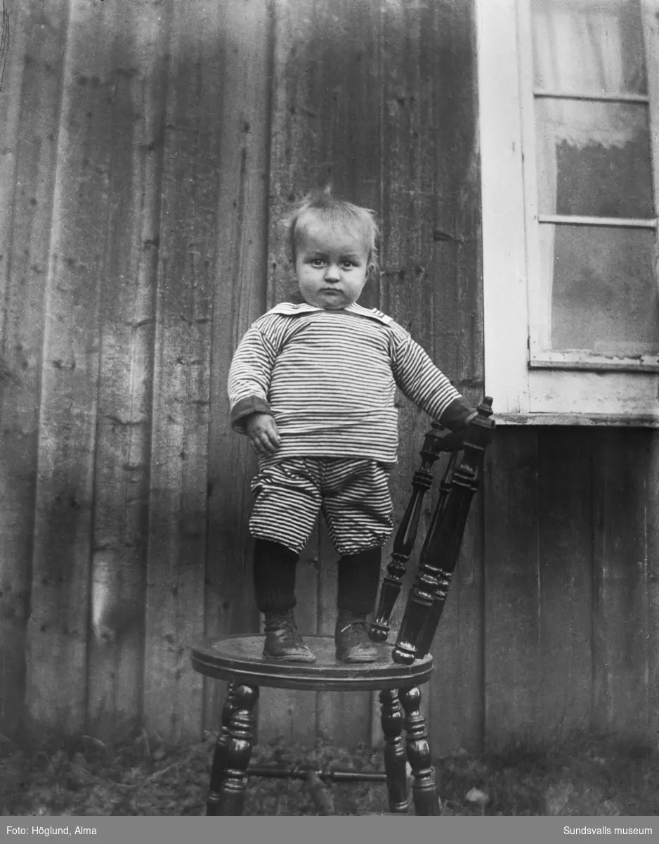 Porträtt av en liten pojke som står på en stol invid en husvägg.